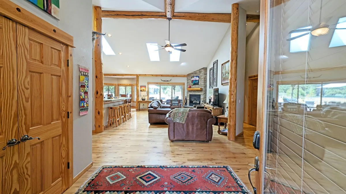 A wide view of the living area featuring exposed wooden beams, a colorful area rug, and expansive windows that flood the space with light. The arrangement suggests a flow between living and dining spaces, with the fireplace as a visual anchor. The scene exudes warmth and a welcoming ambiance for gatherings.