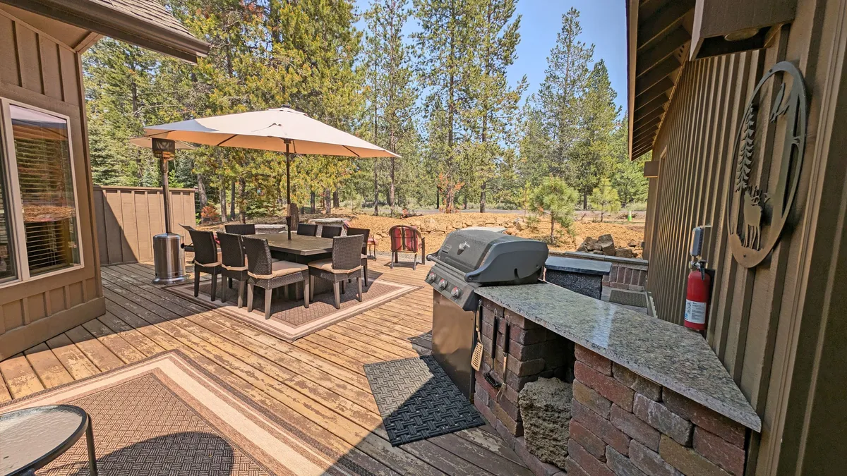 An inviting outdoor space with a large dining table, barbecue grill, and umbrella shaded by surrounding trees. The area provides ample room for gatherings, meals, and enjoying the fresh air, making it a perfect spot for families and their furry companions to relax together.