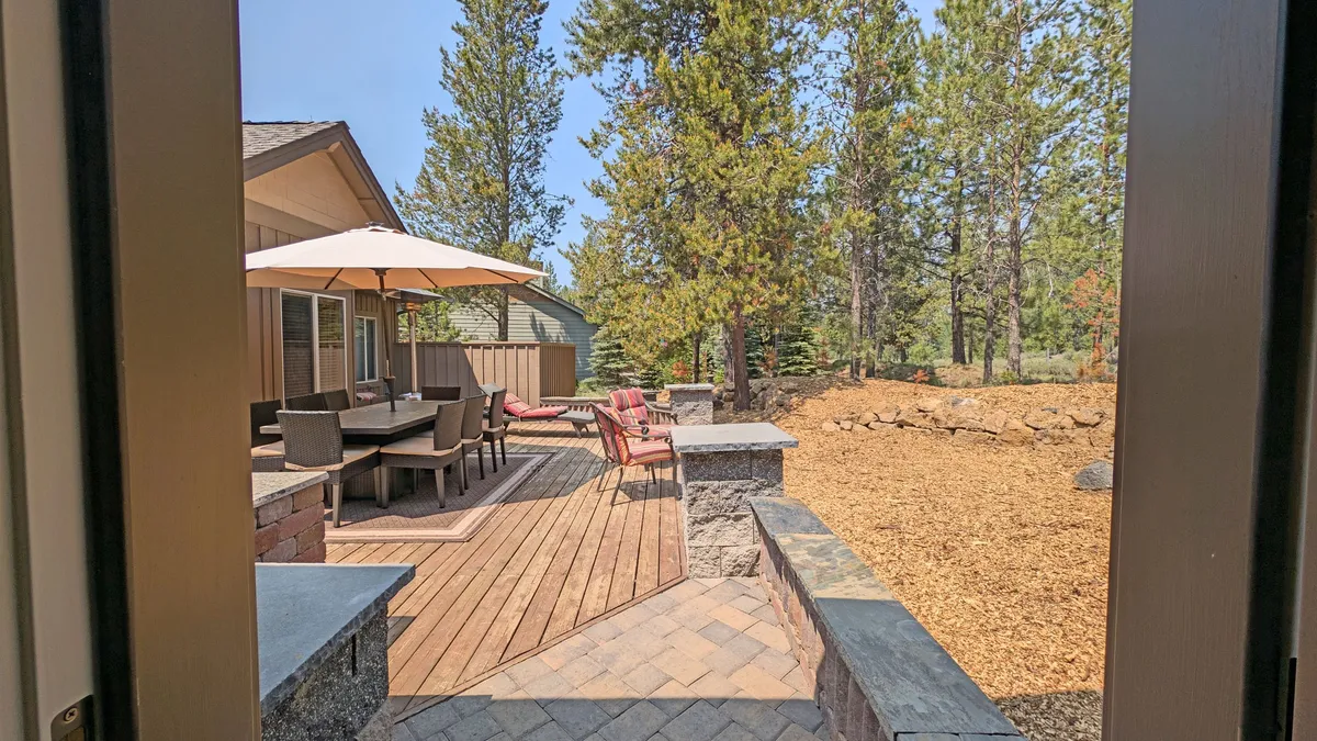 A spacious outdoor dining area nestled among tall trees, featuring a large dining table with an umbrella, surrounded by comfortable seating. A winding pathway leads through the landscaped setting, creating a sense of openness and connection to nature. The area is ideal for relaxing, dining, and socializing after a day of adventure on the trails.