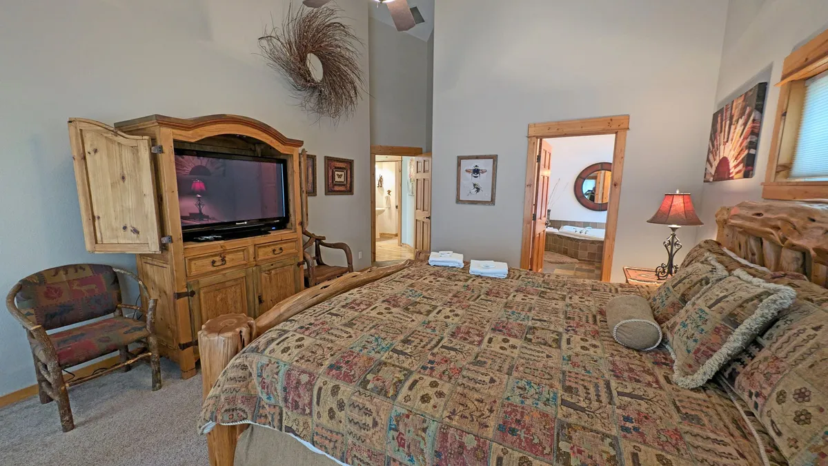 A cozy, inviting bedroom furnished with a large bed with patterned bedding, a wooden armoire with a TV, a decorative chair, and a bedside lamp. The room radiates warmth and offers a perfect spot for quiet relaxation or movie nights in bed.