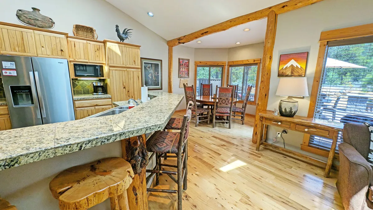 An elegant open-plan kitchen and dining area, highlighted by a granite kitchen island with wooden bar stools. Large windows bathe the space in natural light, and the rustic dining area with unique chairs and visible artwork sets the stage for memorable family meals. The seamless connection between kitchen and dining underscores communal living.