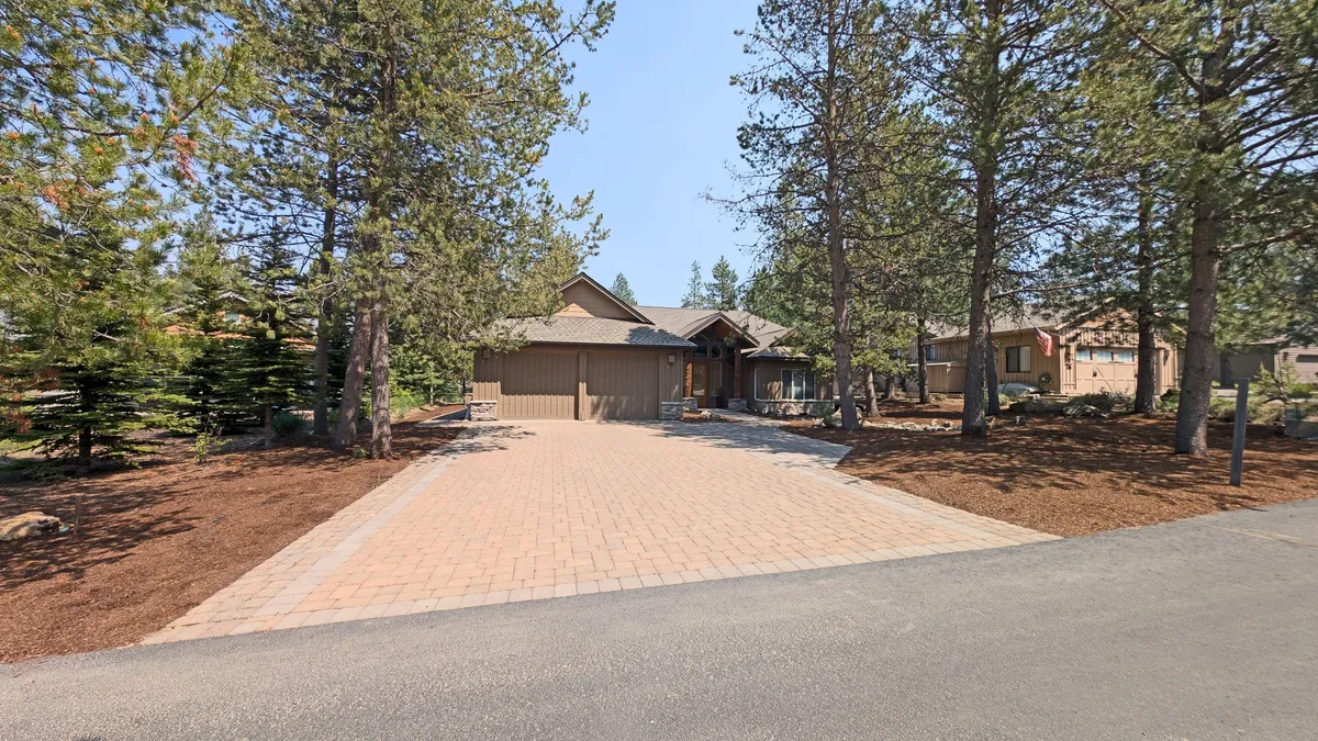 A front exterior view of the home, featuring a welcoming paved driveway, front porch, and tall trees surrounding the property. The image communicates a sense of arrival, the promise of adventure, and a warm, inviting entry for families and groups.