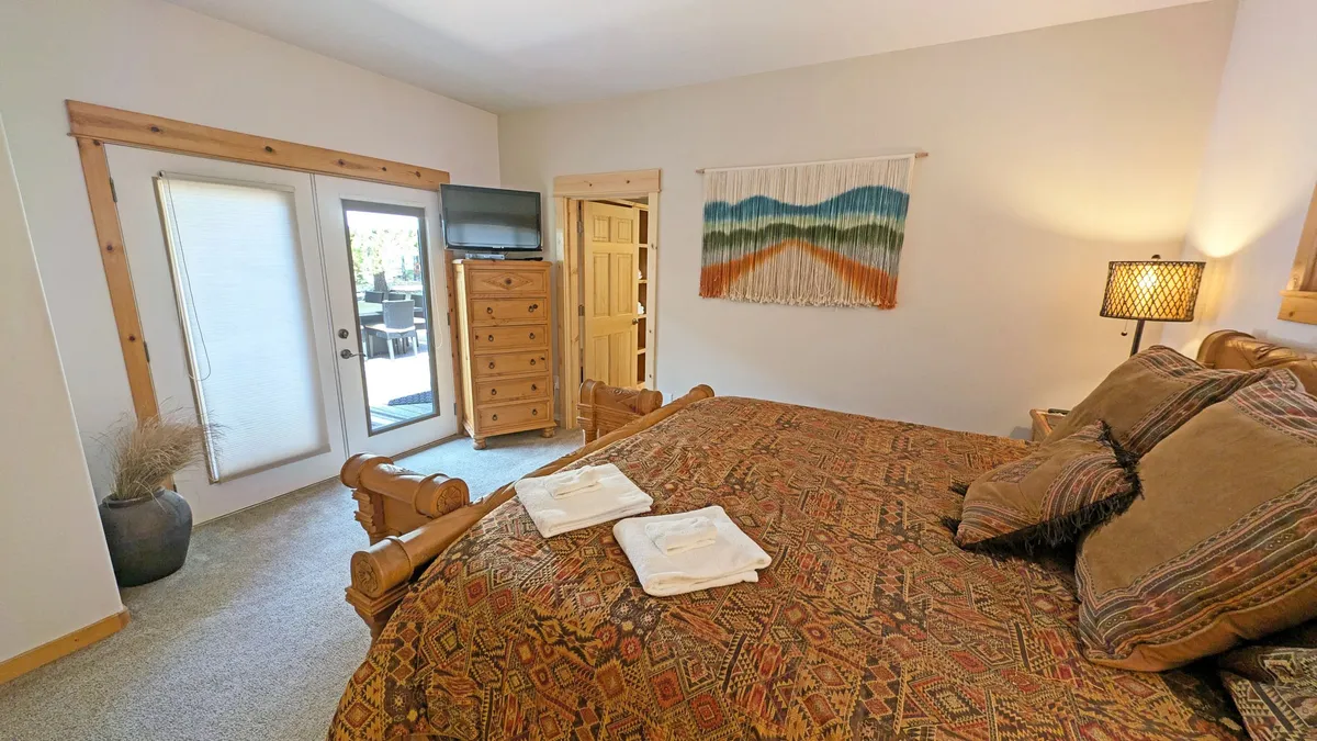 This image features a welcoming bedroom with a bed dressed in patterned bedding, a wooden dresser, wall hanging, and a warm lamp. Natural light filters into the room, creating an atmosphere of comfort and relaxation.