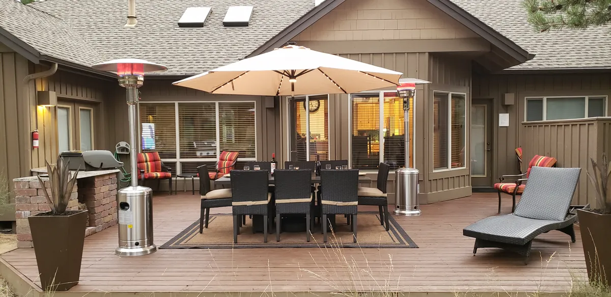 An inviting backyard space featuring a large dining table with chairs beneath a generous umbrella, an outdoor heater for chilly evenings, a lounge chair for relaxation, and a brick grill—all surrounded by the open air. The scene conveys opportunities for post-adventure relaxation and social gatherings.