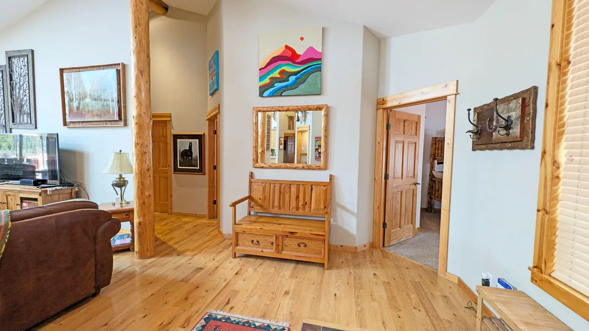 An inviting entryway with a wooden bench, colorful wall art, a large mirror, wooden beams, and a plaid rug. Designed for comfort and practicality, this space is ideal for stowing gear and transitioning from outdoor activities to indoor relaxation.