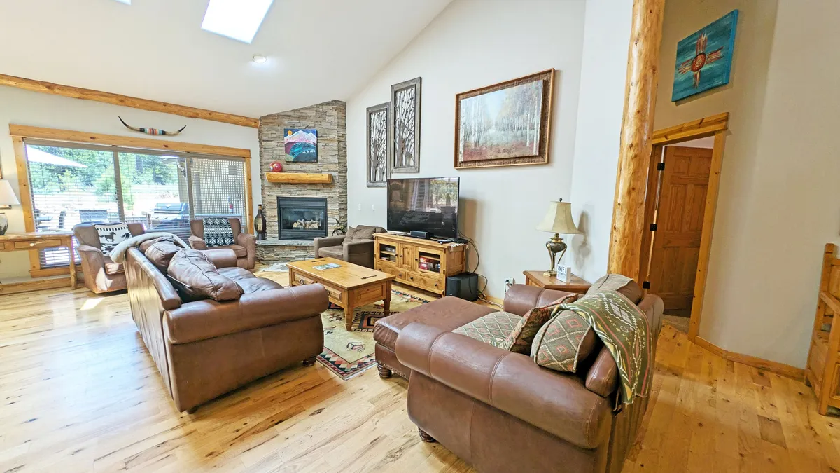 The image showcases a spacious, inviting living room with a plush sofa set arranged around a cozy fireplace, accented by tasteful artwork and bathed in natural light from large windows. The wooden coffee table and expansive layout suggest a perfect setting for group relaxation or gatherings.